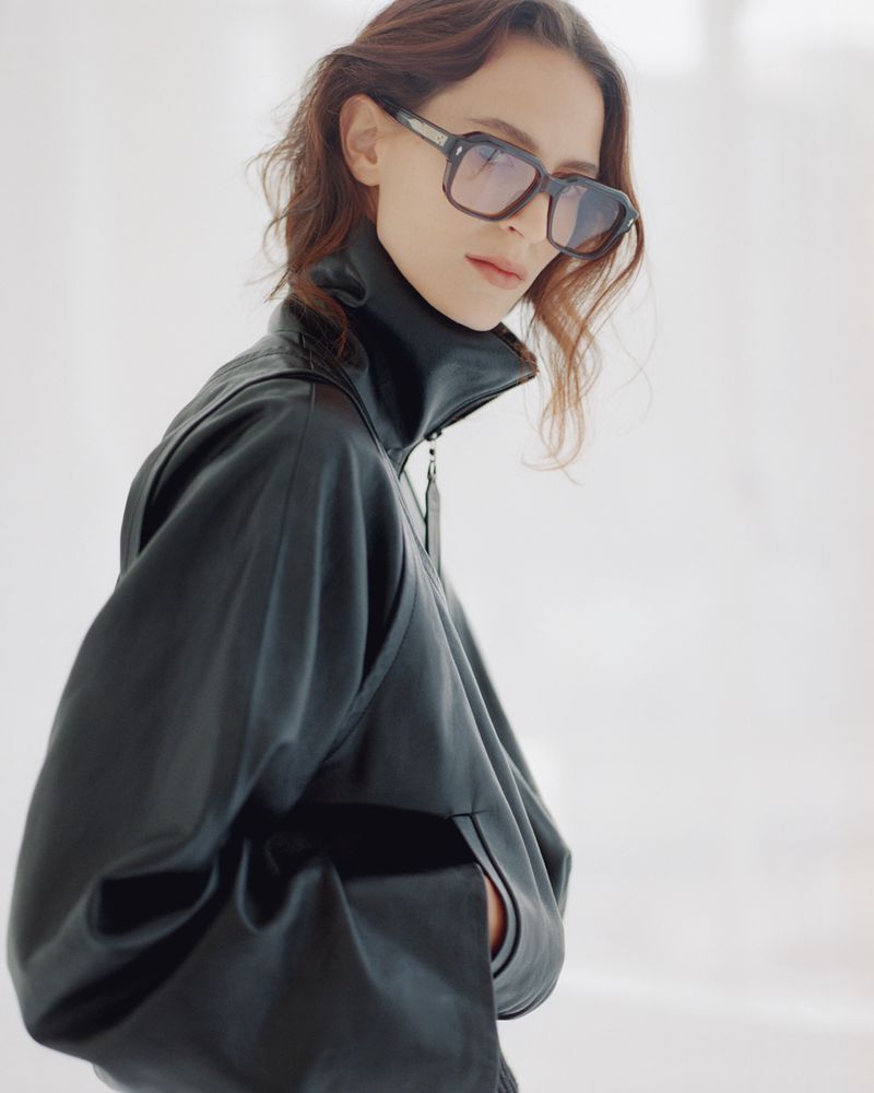 Adult female in a leather jacket and stylish glasses, indoors against a bright white background, fashion-forward and modern.