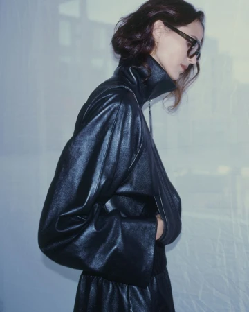 Brunette woman in a black leather jacket and skirt, wearing eyeglasses, posing in front of a bright, neutral background with window light.