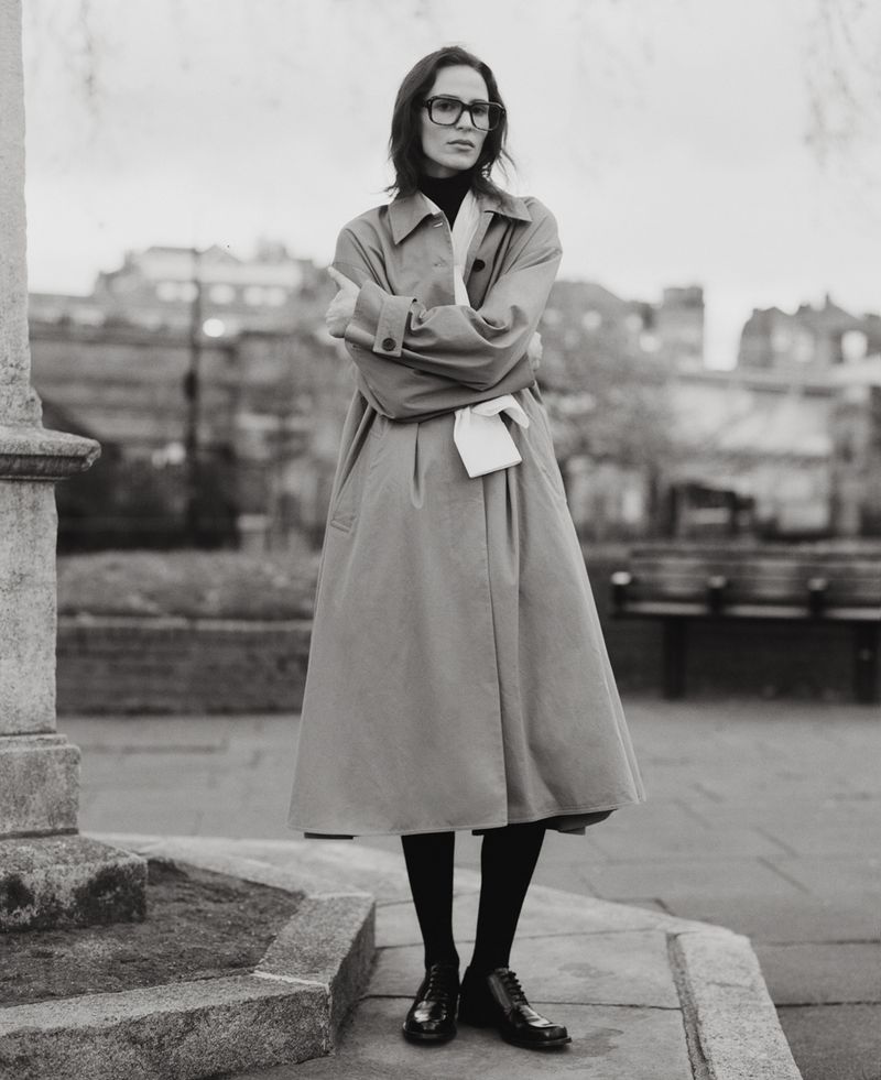 Elegant woman in a tan trench coat stands outdoors with arms crossed, wearing glasses and black tights, in a monochrome fashion portrait.