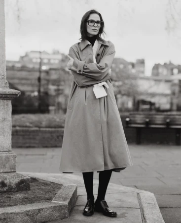 Elegant woman in a tan trench coat stands outdoors with arms crossed, wearing glasses and black tights, in a monochrome fashion portrait.