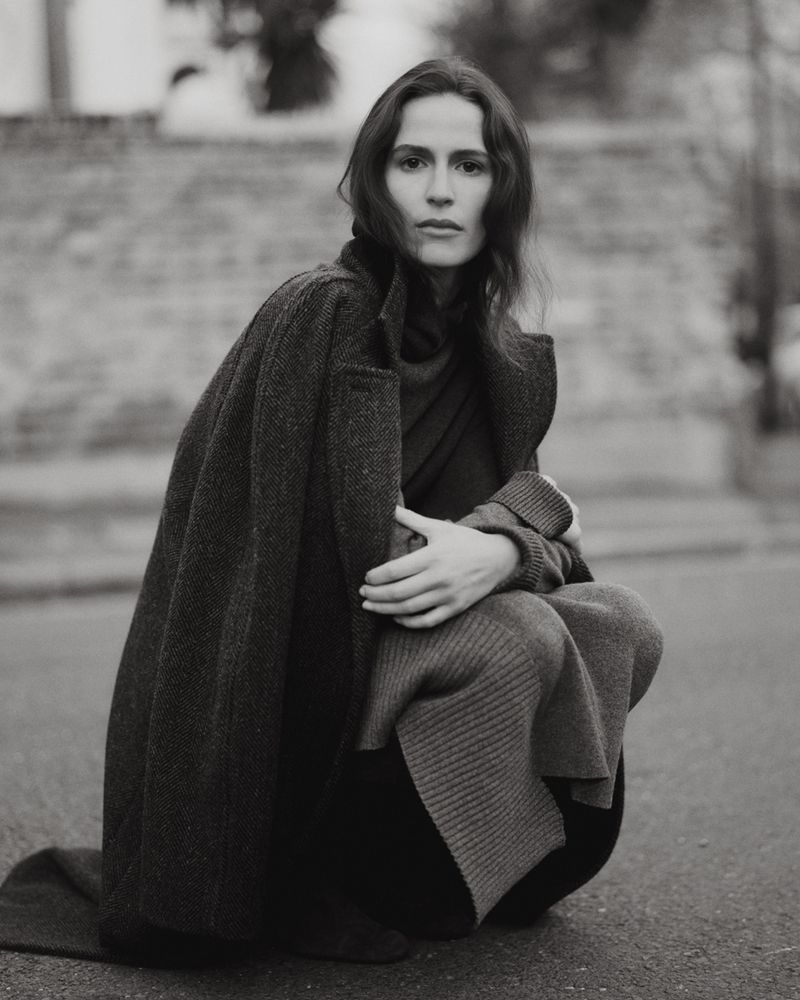 Adult woman with a neutral facial expression squats on a paved surface wearing a long, dark wool coat with a sweater and dark pants.