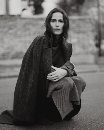 Adult woman with a neutral facial expression squats on a paved surface wearing a long, dark wool coat with a sweater and dark pants.