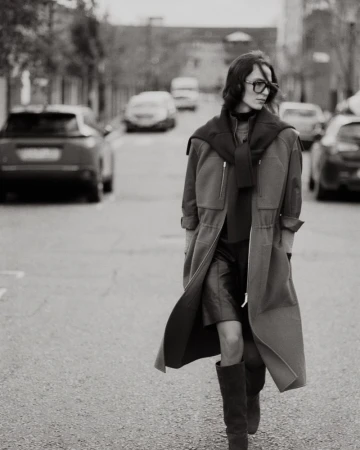 Model Jeanne Cadieu walking down a city street wearing a long open coat, draped sweater, glasses, and tall boots in black and white.