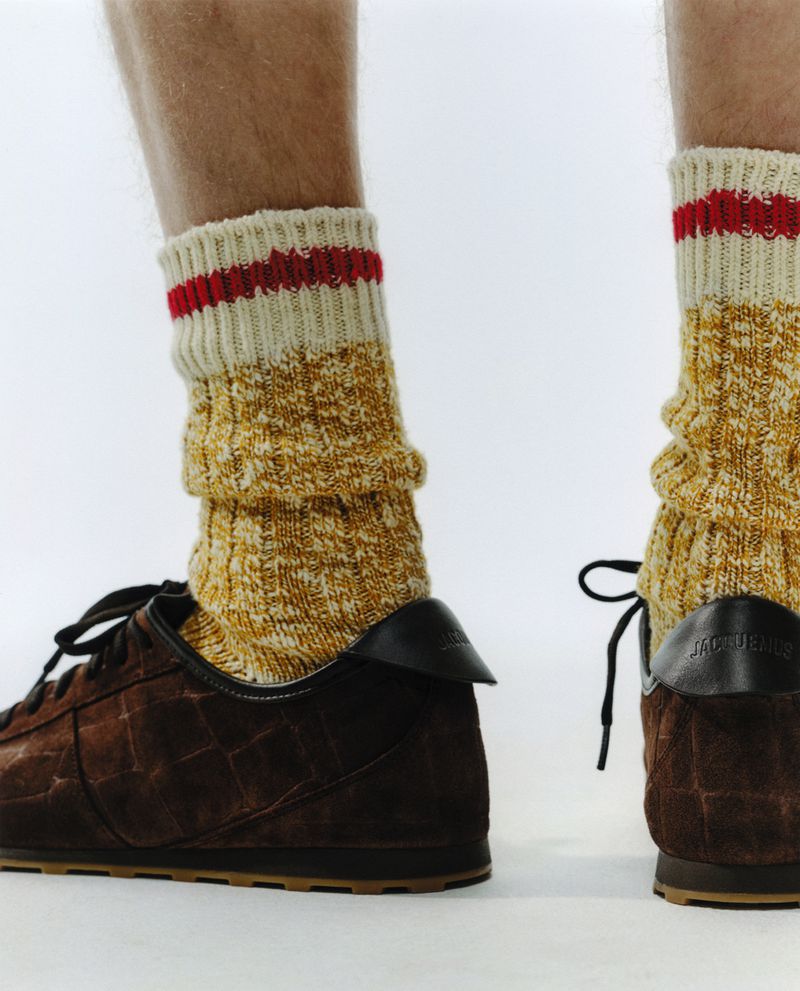 Close-up of a person wearing textured yellow socks with red stripes and brown Jacquemus crocodile print leather sneakers on white backdrop.