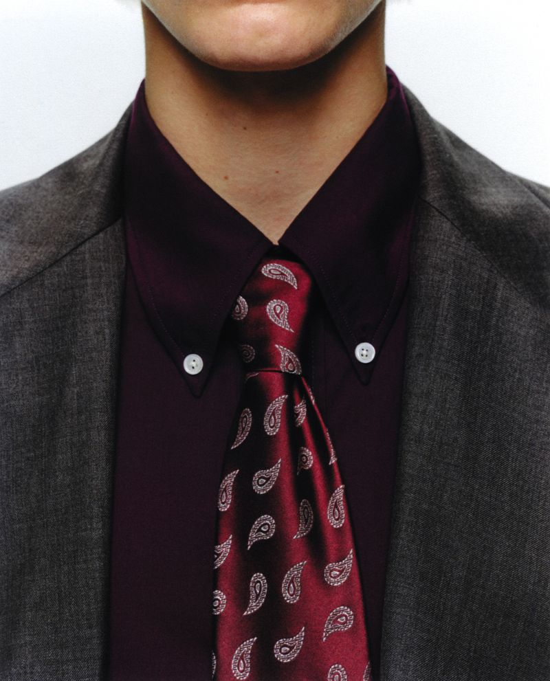 Close-up studio shot of a light-skinned adult wearing a grey suit jacket, dark red button-down shirt, and patterned maroon tie.