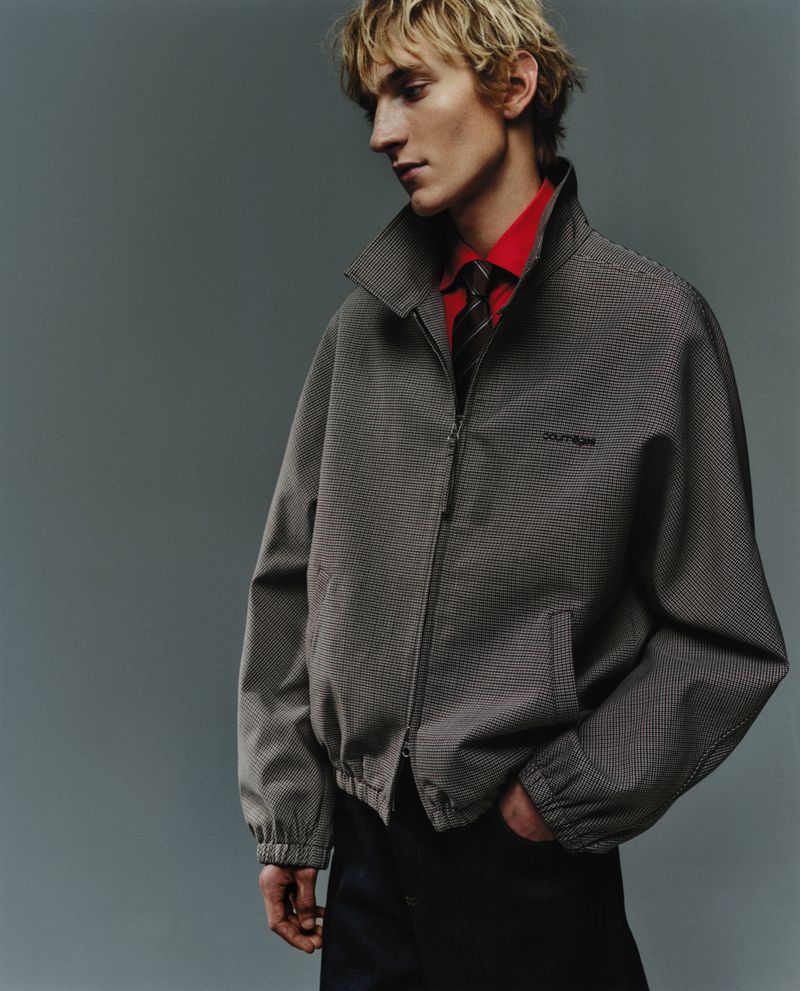 Studio shot of a blonde male model wearing a patterned grey jacket, red collared shirt, and tie, standing against a grey backdrop.