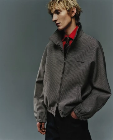 Studio shot of a blonde male model wearing a patterned grey jacket, red collared shirt, and tie, standing against a grey backdrop.