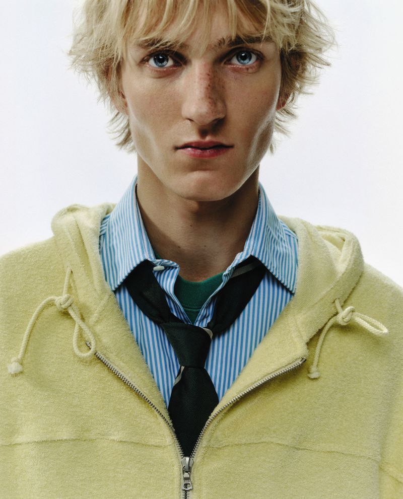 Portrait of a young fair-skinned male model with blond hair and blue eyes wearing a striped shirt, tie, and yellow zip-up hooded jacket.