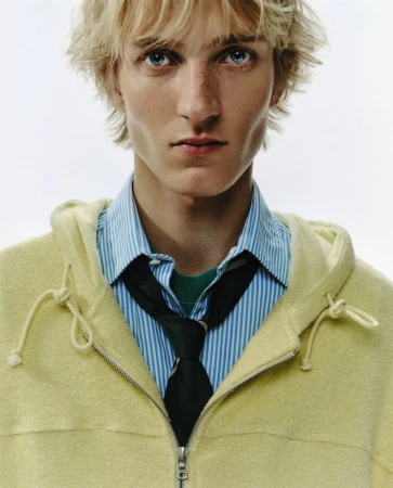 Portrait of a young fair-skinned male model with blond hair and blue eyes wearing a striped shirt, tie, and yellow zip-up hooded jacket.