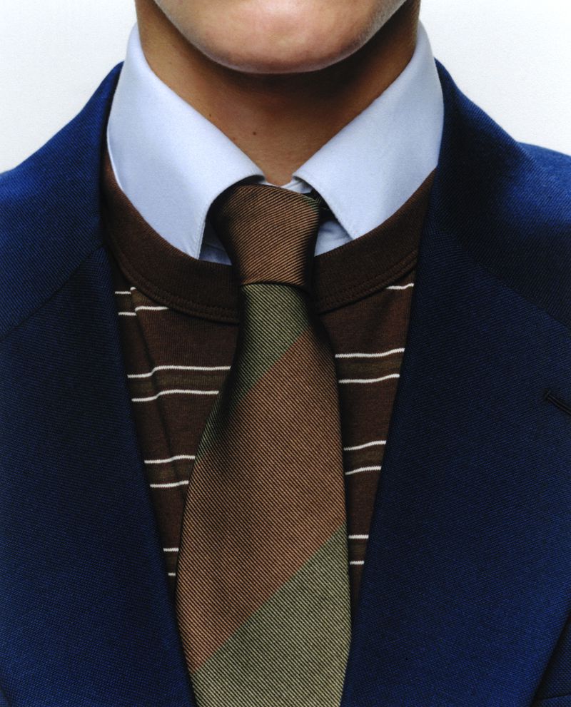 Close-up of a man's torso wearing a blue suit jacket, collared shirt, brown t-shirt, and a diagonally striped brown and green tie.