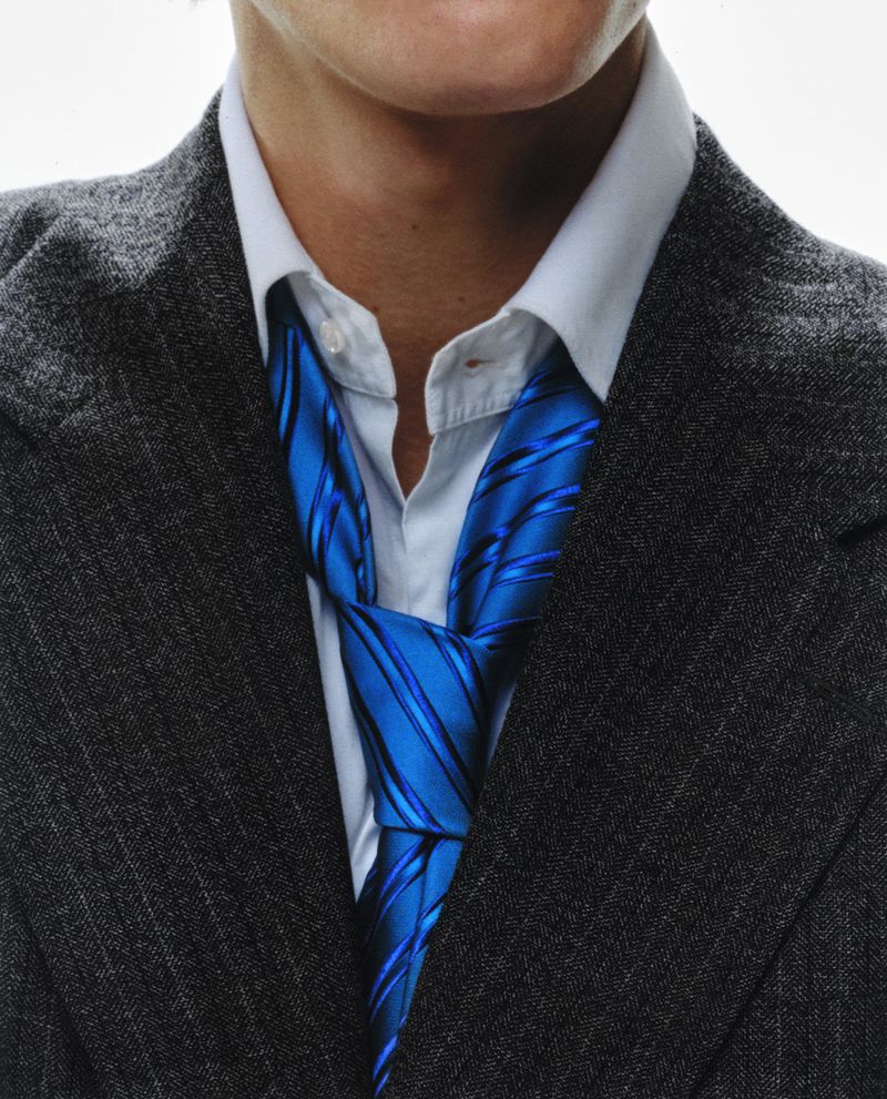 Close-up studio shot of a man wearing a grey suit jacket, a white dress shirt and a knotted blue striped tie, illuminated by studio lighting.