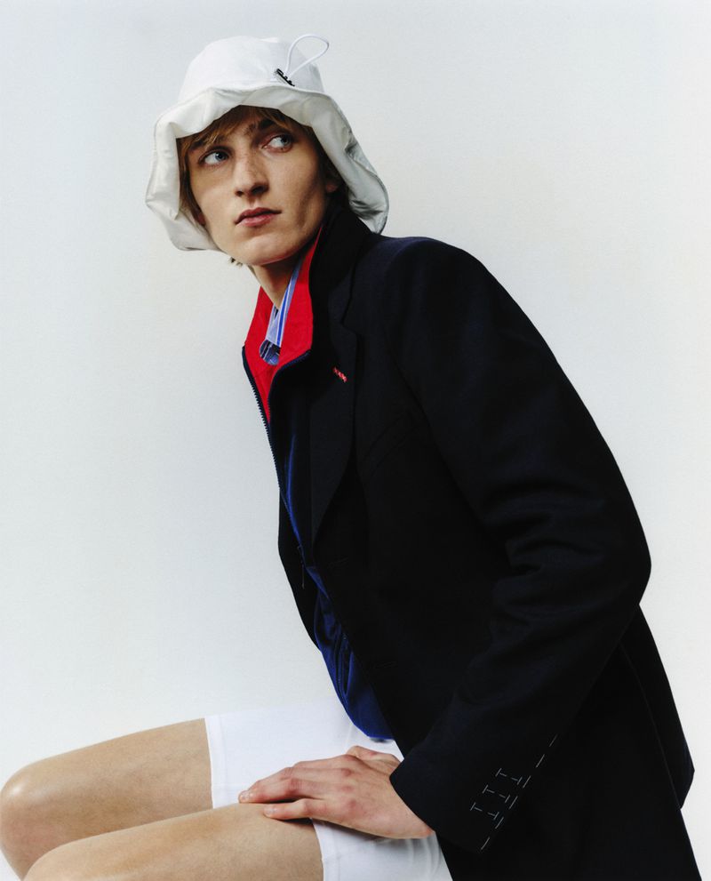 Male fashion model wearing a navy blazer, striped shirt, and white bucket hat posing in a white studio environment for a fashion editorial.