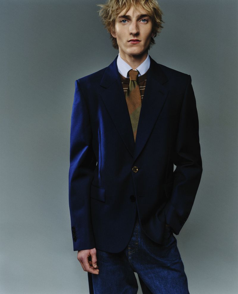 Portrait of a blond male model wearing a navy blazer, patterned tie, and jeans against a grey background in a studio setting.