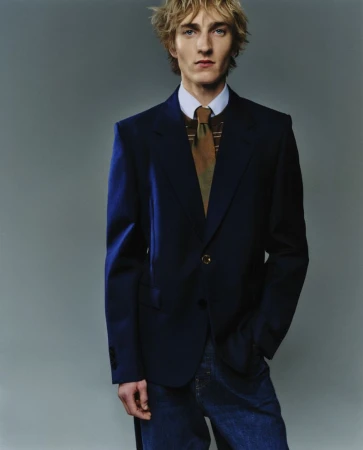Portrait of a blond male model wearing a navy blazer, patterned tie, and jeans against a grey background in a studio setting.