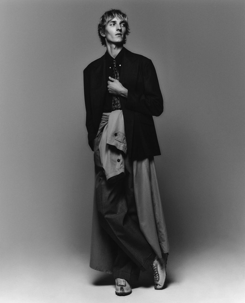 Black and white portrait of a slender adult male fashion model in a blazer, button-down shirt, and deconstructed pants in a photo studio.