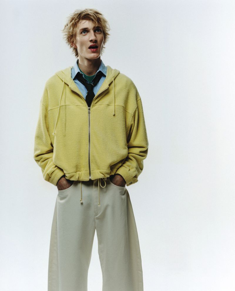 A blond male model gazes upward in a studio shot, wearing a yellow hooded sweatshirt, light blue shirt, dark tie, and baggy white pants.