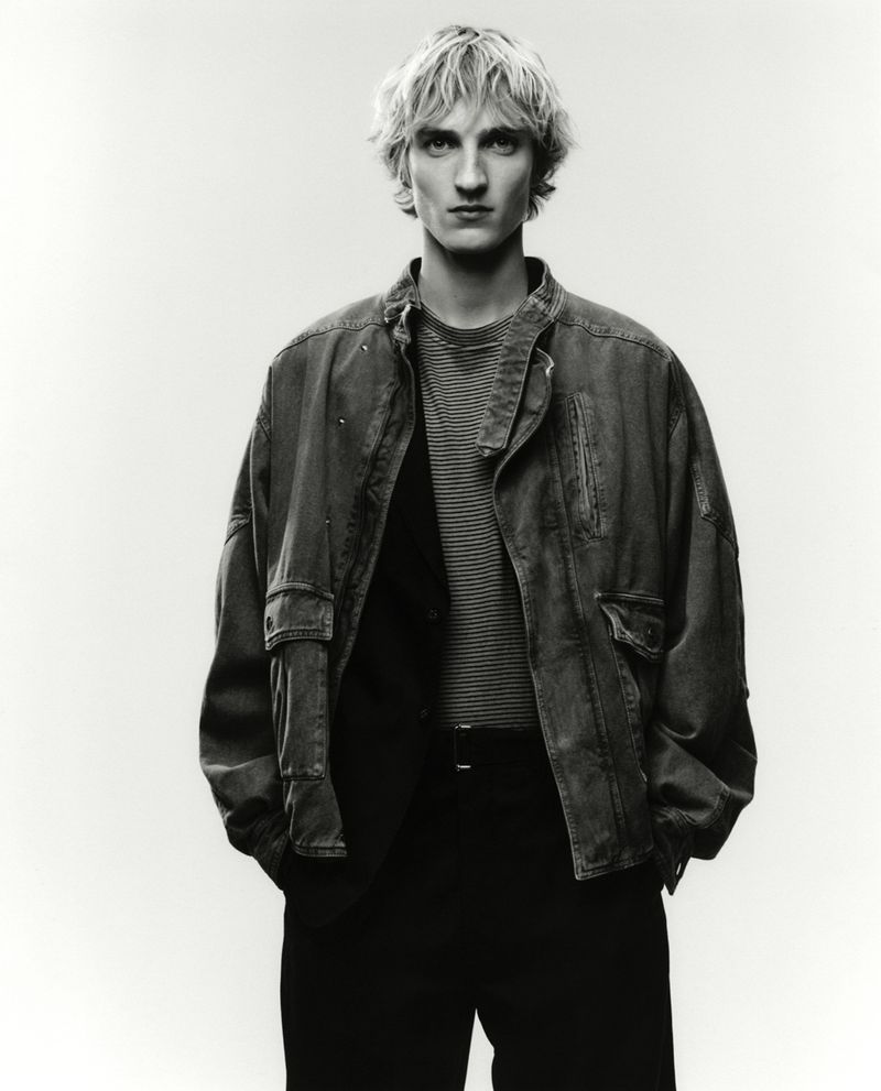 A blond male model poses in a gray jacket and striped shirt against a white backdrop, fashion shoot, studio lighting.