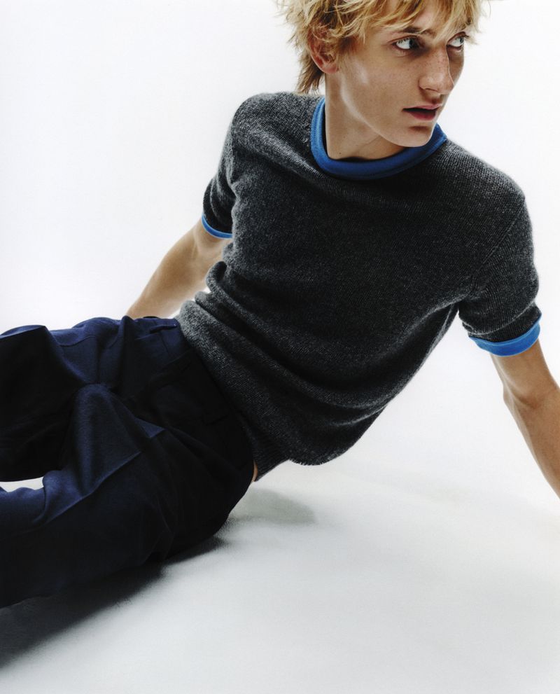 Young adult male with blond hair in a grey short-sleeved shirt with blue trim and navy pants poses in front of a bright white background.