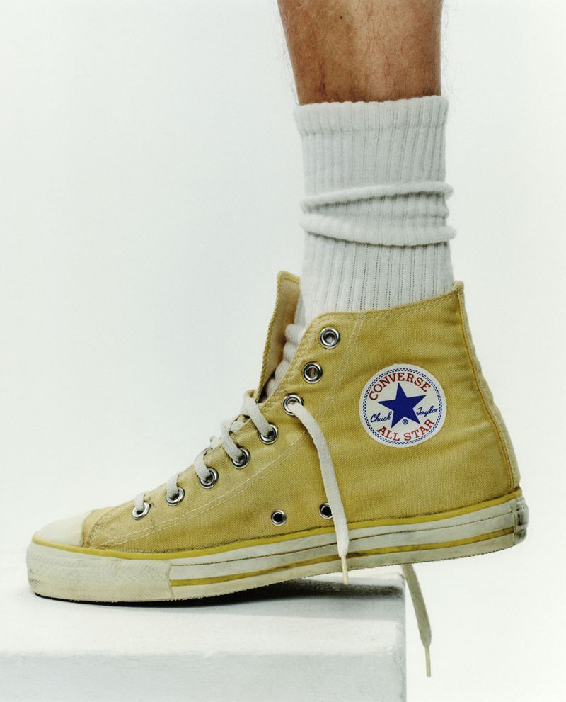 Tan Converse high-top sneaker on a white platform with a person wearing a white sock; product shot for fashion and style.