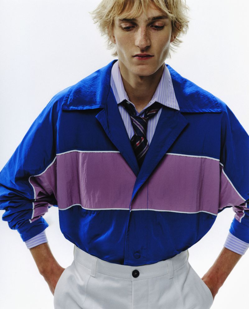 Fashion portrait of a young adult male wearing a blue, pink, and white jacket, striped shirt, and tie against a white backdrop.