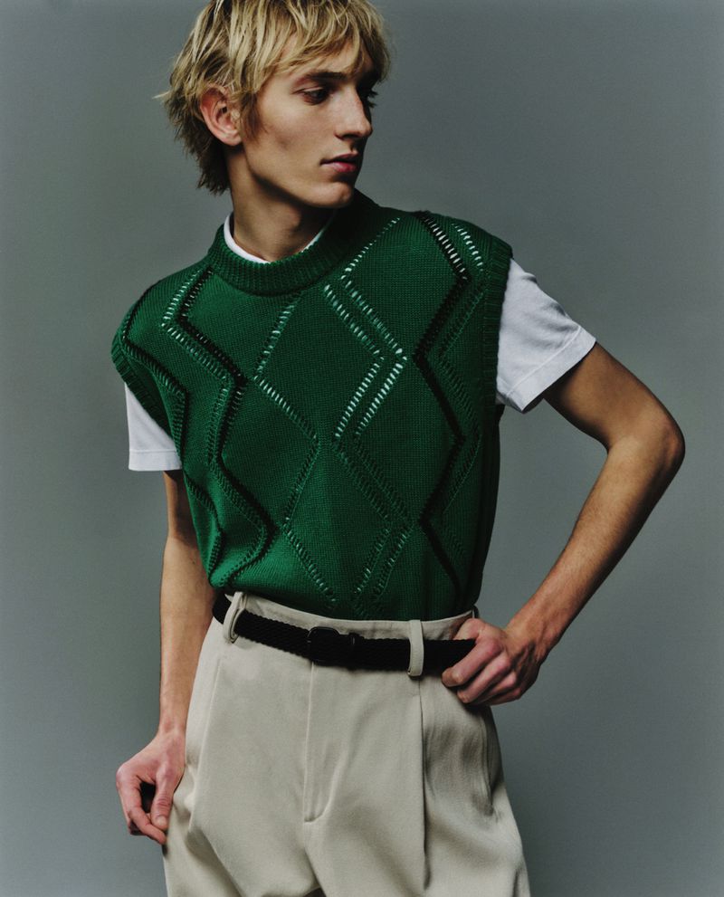 Blond-haired man poses in a green argyle sweater vest with khaki pants and a black belt against a grey studio backdrop in a fashion shot.