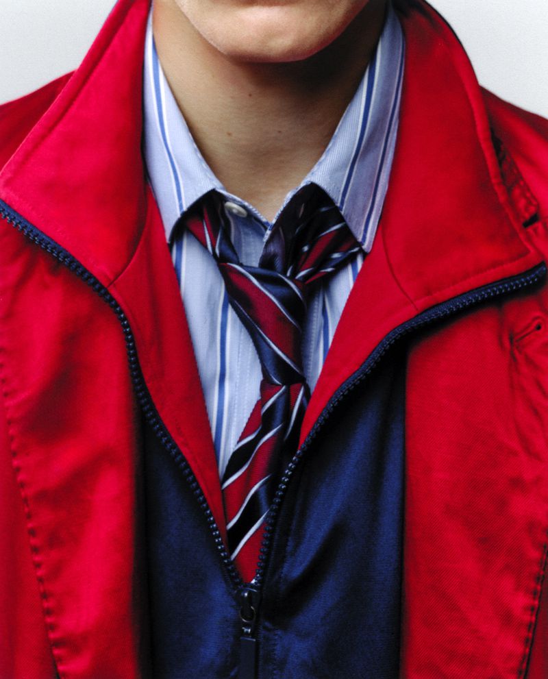 Closeup of a person wearing a striped dress shirt, navy vest, and a loosely knotted tie, with a bright red, unzipped jacket on top.