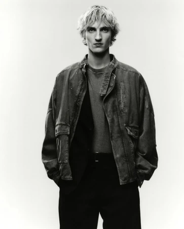 Model Pierrick Grégoire posed in an oversized utility jacket, striped shirt, and black trousers against a bright studio background.