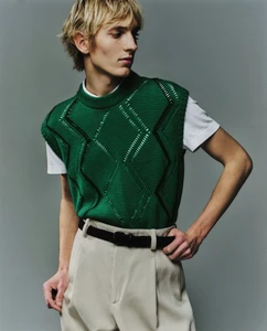 Model Pierrick Grégoire wearing an emerald green patterned knit vest, white t-shirt, and beige pleated trousers against a grey backdrop.