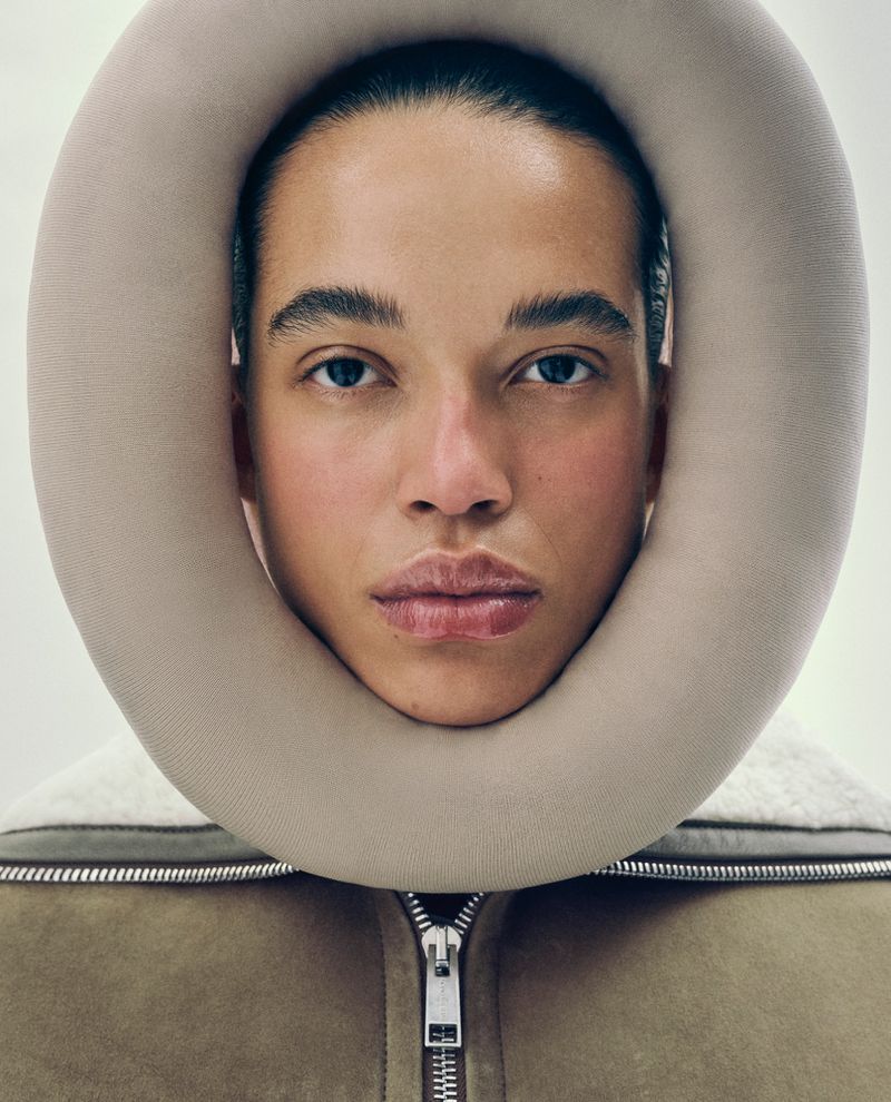 Close-up studio portrait of a fair-skinned person with dark hair wearing a beige jacket and a large neck support pillow.