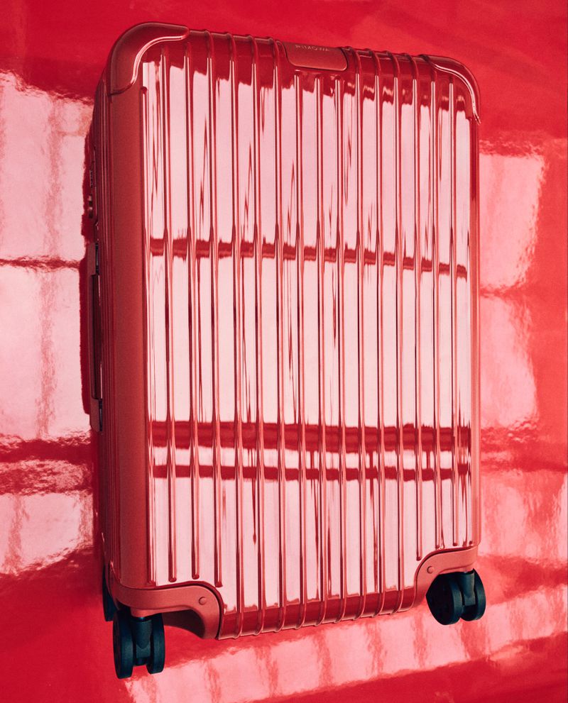 A red metallic suitcase with wheels stands against a glossy red background, reflecting light in a minimalist travel product shot.