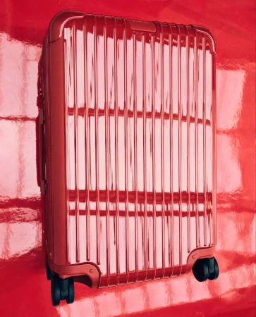 A red metallic suitcase with wheels stands against a glossy red background, reflecting light in a minimalist travel product shot.