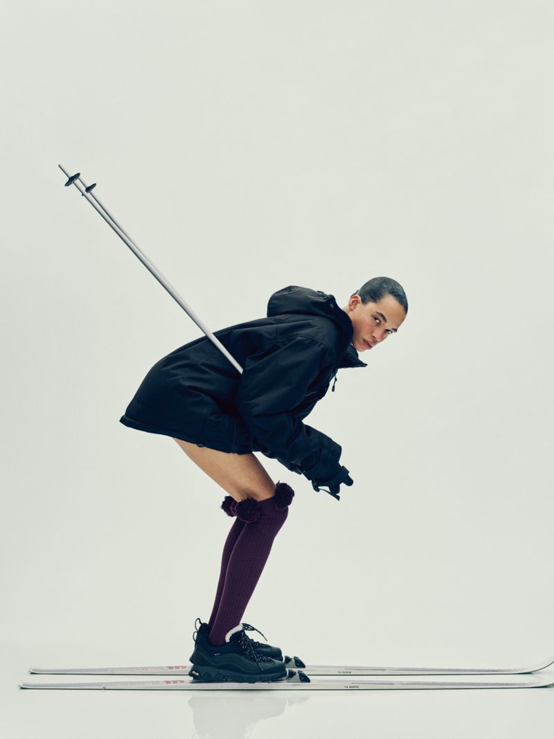 Fashion shot of a young adult in a dark jacket and high socks posing on skis in a studio with ski poles. Bright, minimalist background.