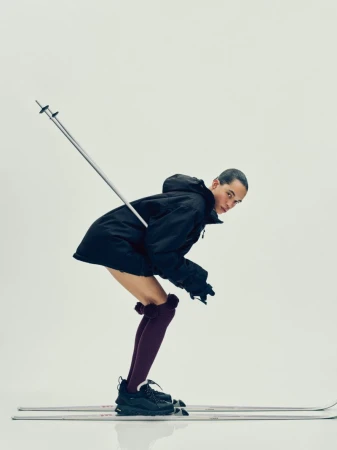 Fashion shot of a young adult in a dark jacket and high socks posing on skis in a studio with ski poles. Bright, minimalist background.