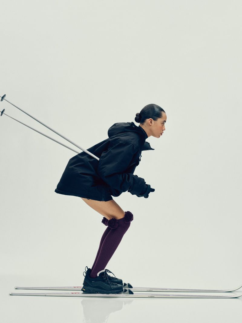 Fashionable adult female on skis in a studio. She wears a black coat, black shoes, and maroon stockings with pom poms against a white backdrop.