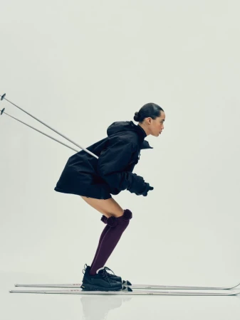 Fashionable adult female on skis in a studio. She wears a black coat, black shoes, and maroon stockings with pom poms against a white backdrop.