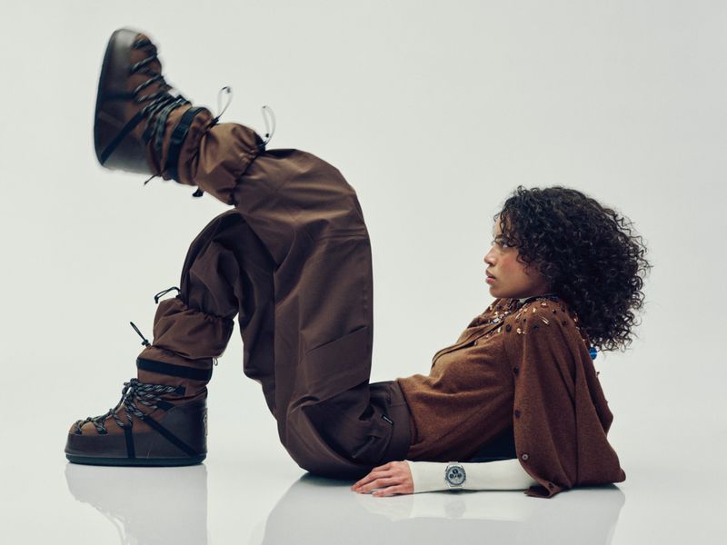 A person with curly hair is posing in brown snow boots and matching brown pants and a jacket while lying on a white surface.