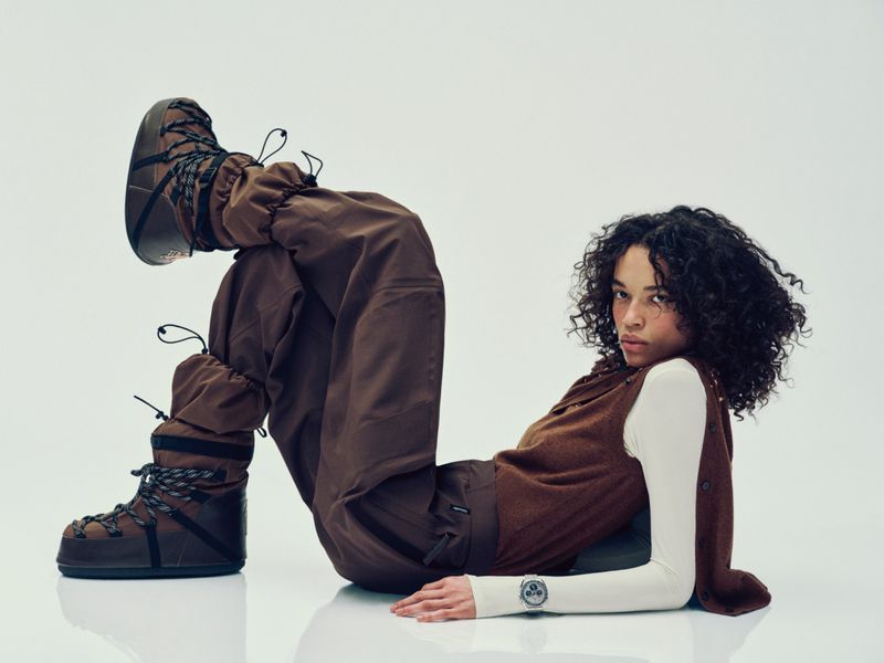 A person with curly hair poses in brown snow boots, cargo pants, vest, and long-sleeve shirt on a white backdrop in a studio setting.