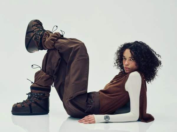 A person with curly hair poses in brown snow boots, cargo pants, vest, and long-sleeve shirt on a white backdrop in a studio setting.