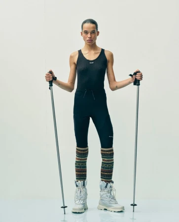 Full shot of androgynous model posing in black athletic wear, leg warmers, and white boots, holding ski poles against a white backdrop.