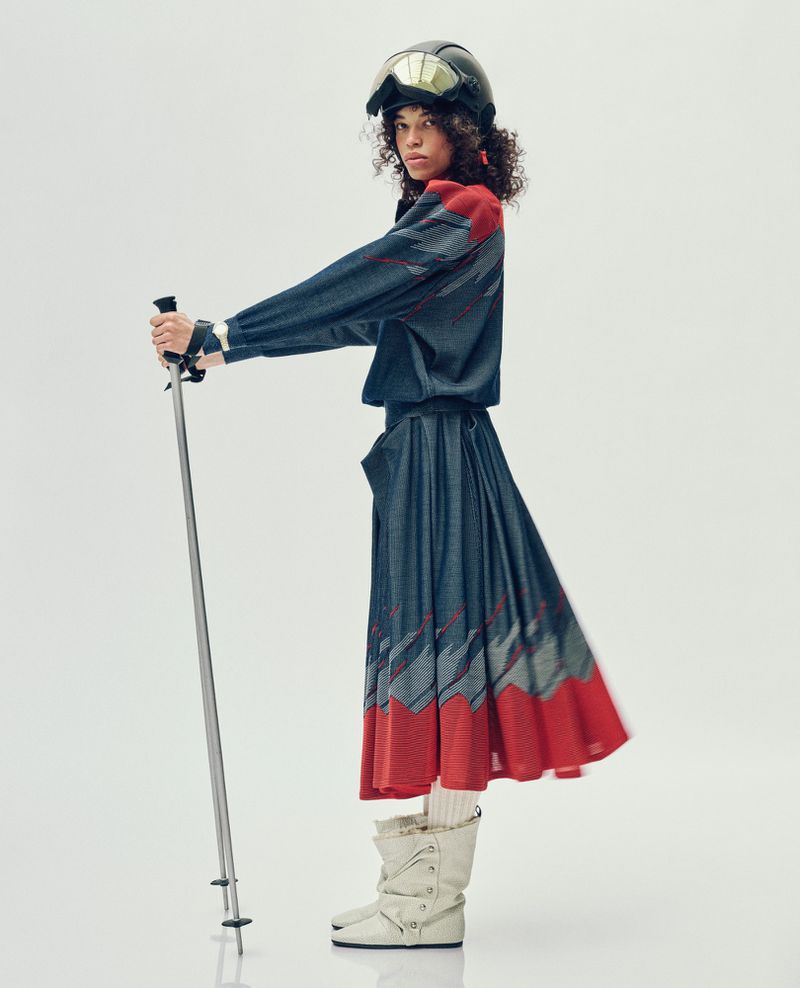Woman in a vintage ski outfit and black helmet poses with ski poles against a white backdrop, showcasing winter sport fashion.