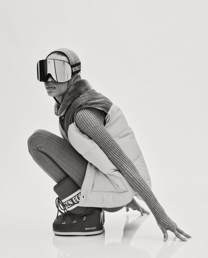 A female model poses in ski goggles, a beanie, a puffer vest, leggings, and Moon Boots in a white photo studio setting.