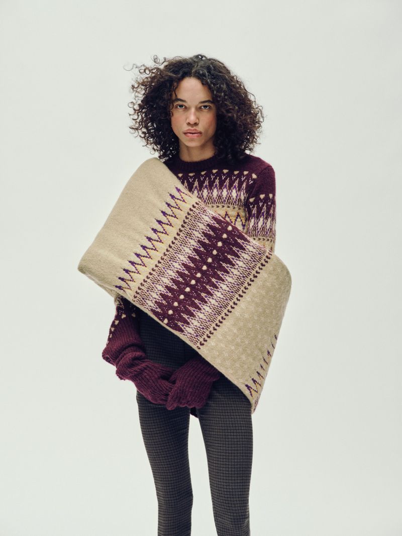 A young woman with curly hair poses indoors, wearing a patterned sweater, gloves, and a patterned beige shawl against a plain background.