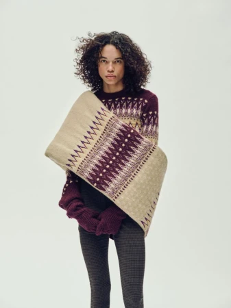 A young woman with curly hair poses indoors, wearing a patterned sweater, gloves, and a patterned beige shawl against a plain background.
