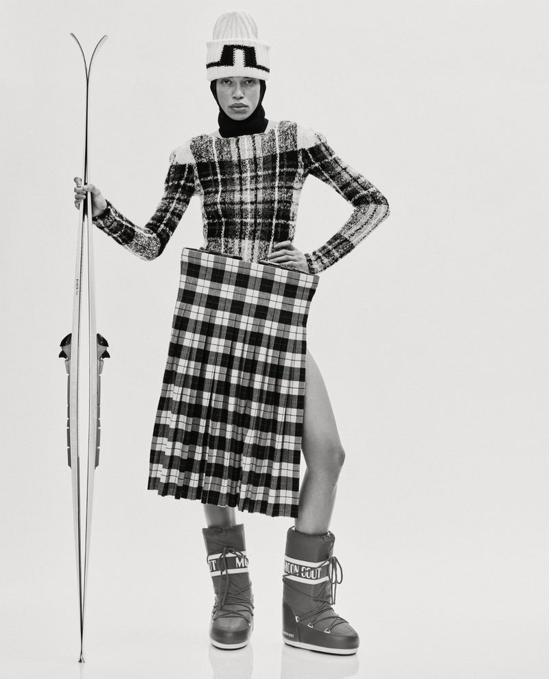 Woman in avant-garde fashion poses in a studio with skis, wearing a plaid skirt, a sweater, winter boots, and a knit hat.