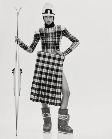 Woman in avant-garde fashion poses in a studio with skis, wearing a plaid skirt, a sweater, winter boots, and a knit hat.