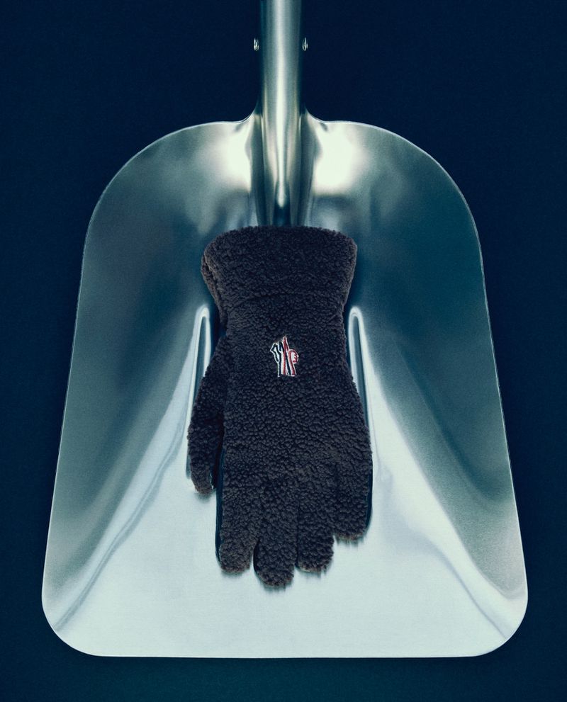 A black glove with a logo sits on a silver metal shovel against a dark blue backdrop in a product-style studio shot.