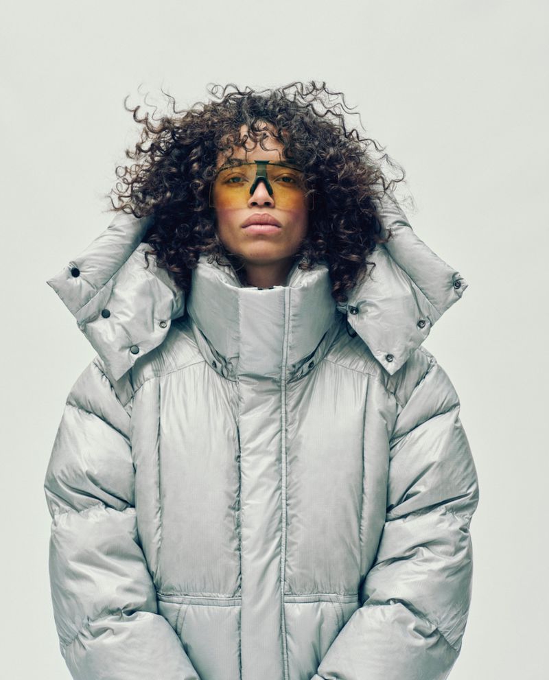 Portrait of a curly-haired adult female wearing stylish yellow sunglasses and a silver puffy hooded jacket against a neutral background.