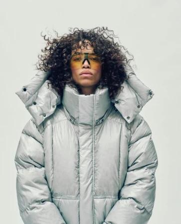 Portrait of a curly-haired adult female wearing stylish yellow sunglasses and a silver puffy hooded jacket against a neutral background.