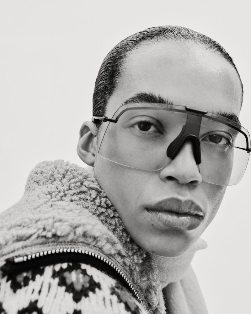 Close-up black and white studio shot of a person wearing sporty sunglasses and a patterned wool jacket against a plain white backdrop.