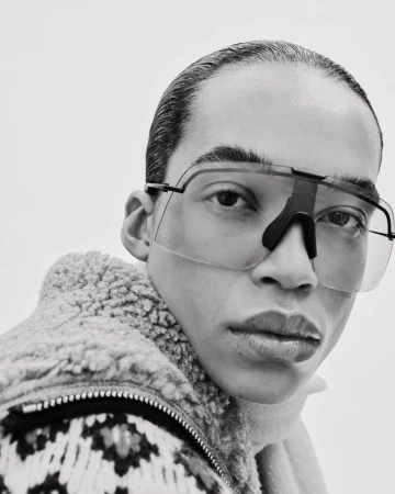 Close-up black and white studio shot of a person wearing sporty sunglasses and a patterned wool jacket against a plain white backdrop.